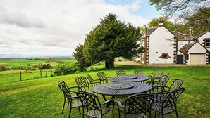 A dining area with a round table and chairs, surrounded by a grassy lawn, with a large tree and a building in the background. - Kinclune House & Cottages - Sleeps 28 (Kirkton of Kingoldrum)