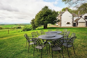 Un comedor con una mesa redonda y sillas, rodeado por un césped, con un árbol grande y un edificio al fondo.