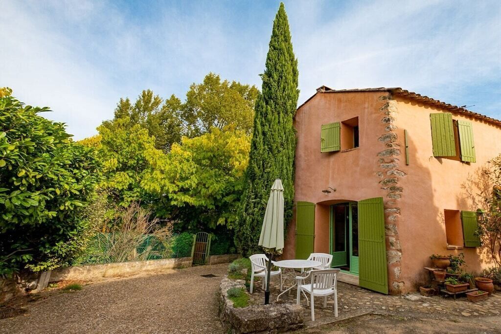 Edifici rústic amb façana de pedra, persianes verdes i un patió amb mobles de jardí.