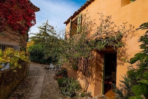 A narrow stone pathway leading to a building with a rustic exterior, surrounded by lush vegetation and a small patio area. - In a quiet location in the Luberon - 219 ROU (Gargas)
