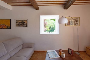 A room with a sofa, a table with a lamp, framed pictures, and a window with a view of greenery. - Holiday home with 4 bedrooms (Roussillon)