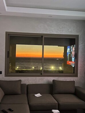 A room with a large window overlooking a sunset over the water, a sofa, and a television screen displaying a news program. - Elegant 3 bed apartment with sea view (Anza)
