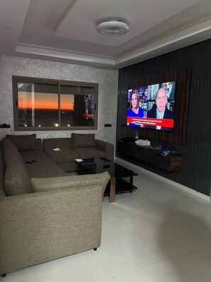 A modern living room with a large flat-screen TV, a sectional sofa, and a large window with a view of the sunset. - Elegant 3 bed apartment with sea view (Anza)