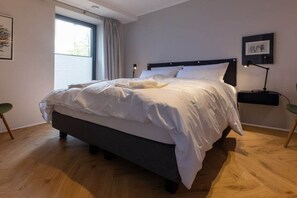 A neatly made bed in a room with a large window, a bedside table with a lamp, and a chair. - Holiday apartment with 1 bedroom (Wildeshausen)