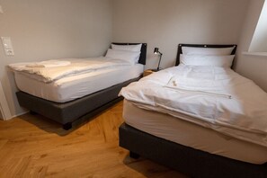 Una habitación con dos camas individuales, cada una con cabecera, mesita de noche y lámpara. El suelo es de madera y las paredes son blancas.