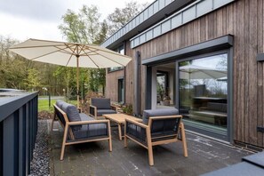 Eine Außenpatio mit Tisch und Stühlen, einem großen Sonnenschirm und einem Holzbau mit Glastür.