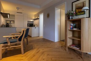 Ein moderner Wohnraum mit Holzboden, einem Essbereich mit Stühlen, einer Küche mit weißen Schränken und einem Bücherregal mit Büchern.