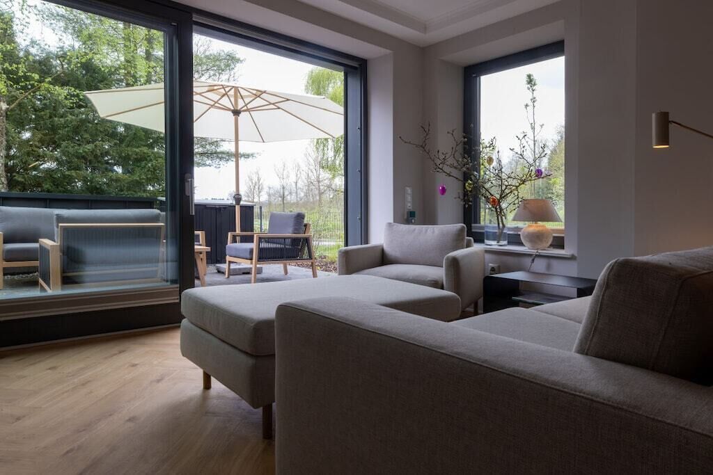 Un salon moderne avec une grande fenêtre, un canapé, un fauteuil et une petite table avec une lampe.
