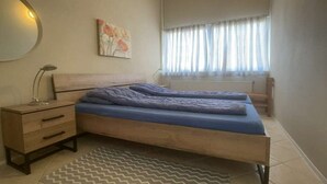A bedroom with a bed, bedside table, lamp, and a window with curtains. - Holiday home with 3 bedrooms (Cadzand-Bad)