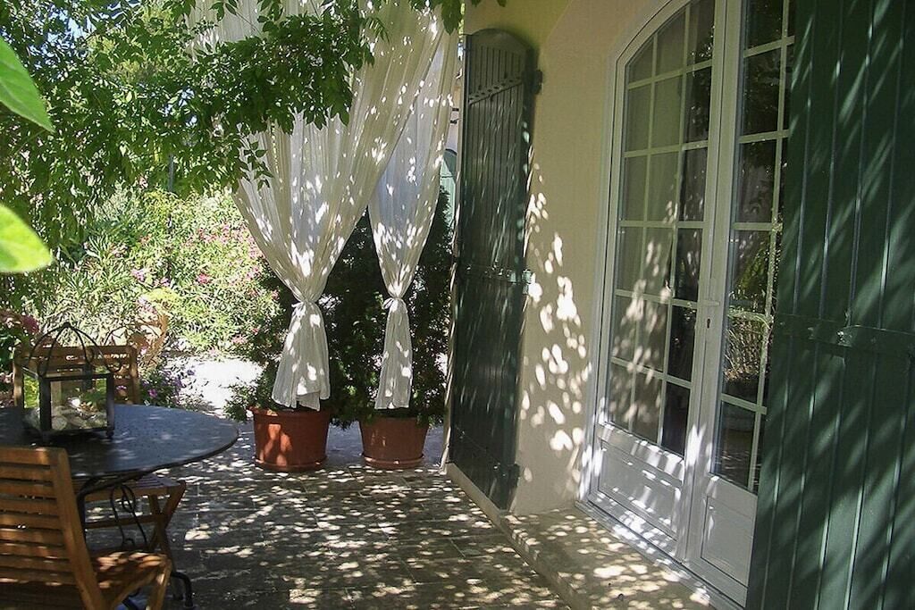 An outdoor patio with a table and chairs, potted plants, and a window with a view of greenery.