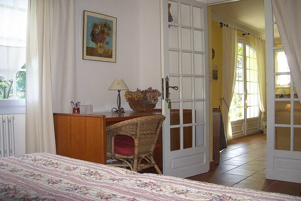 Une chambre à coucher comprenant un lit, un bureau, une chaise en osier, une lampe, un tableau et une porte donnant sur une autre pièce.