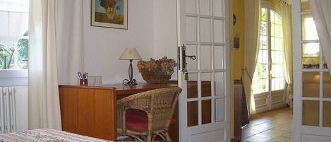 Une chambre à coucher comprenant un lit, un bureau, une chaise en osier, une lampe, un tableau et une porte donnant sur une autre pièce.