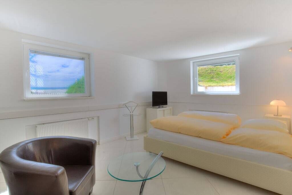 A bedroom with a bed, a TV, a chair, and two windows.