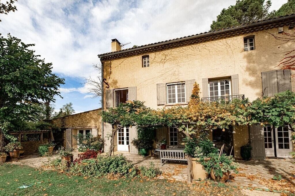 A two-story stone house with a garden, a bench, and a pergola covered with vines.