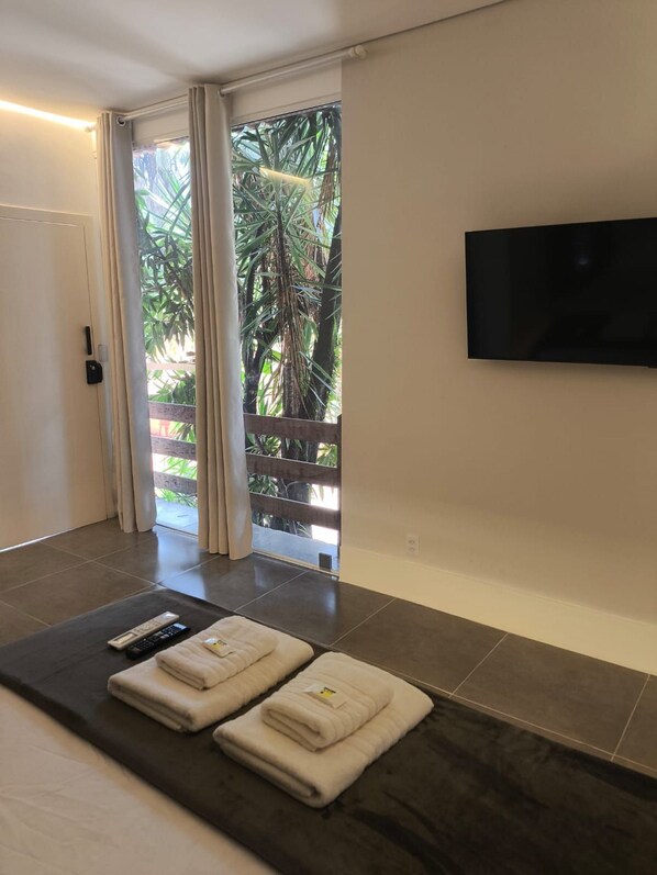 A hotel room with a bed, a flat-screen TV, a balcony with curtains, and a view of greenery. - Gyn Suites Sul (Goiania)