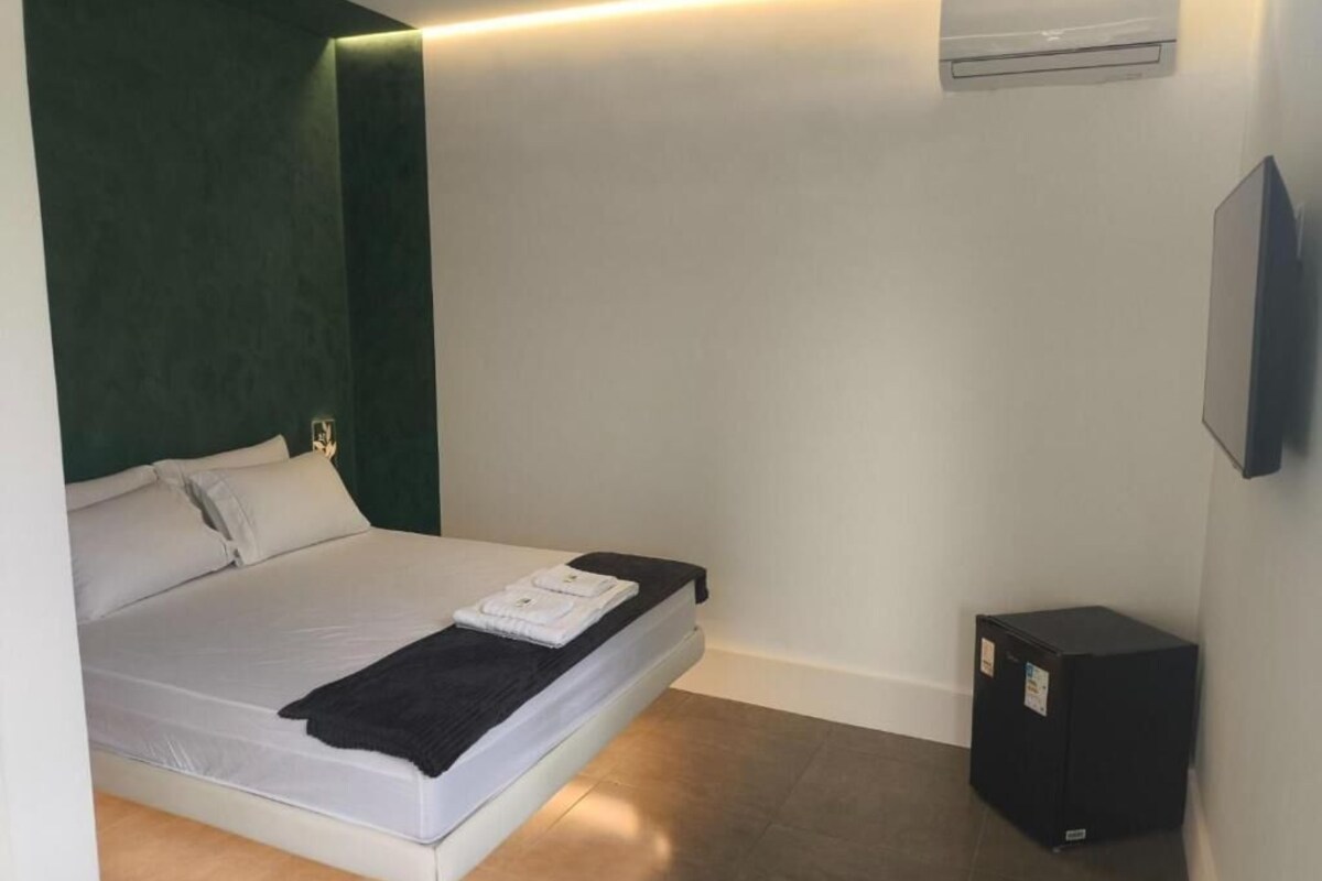 A single bed with a white mattress, a black blanket, and a pillow. There is a small black speaker on the floor.