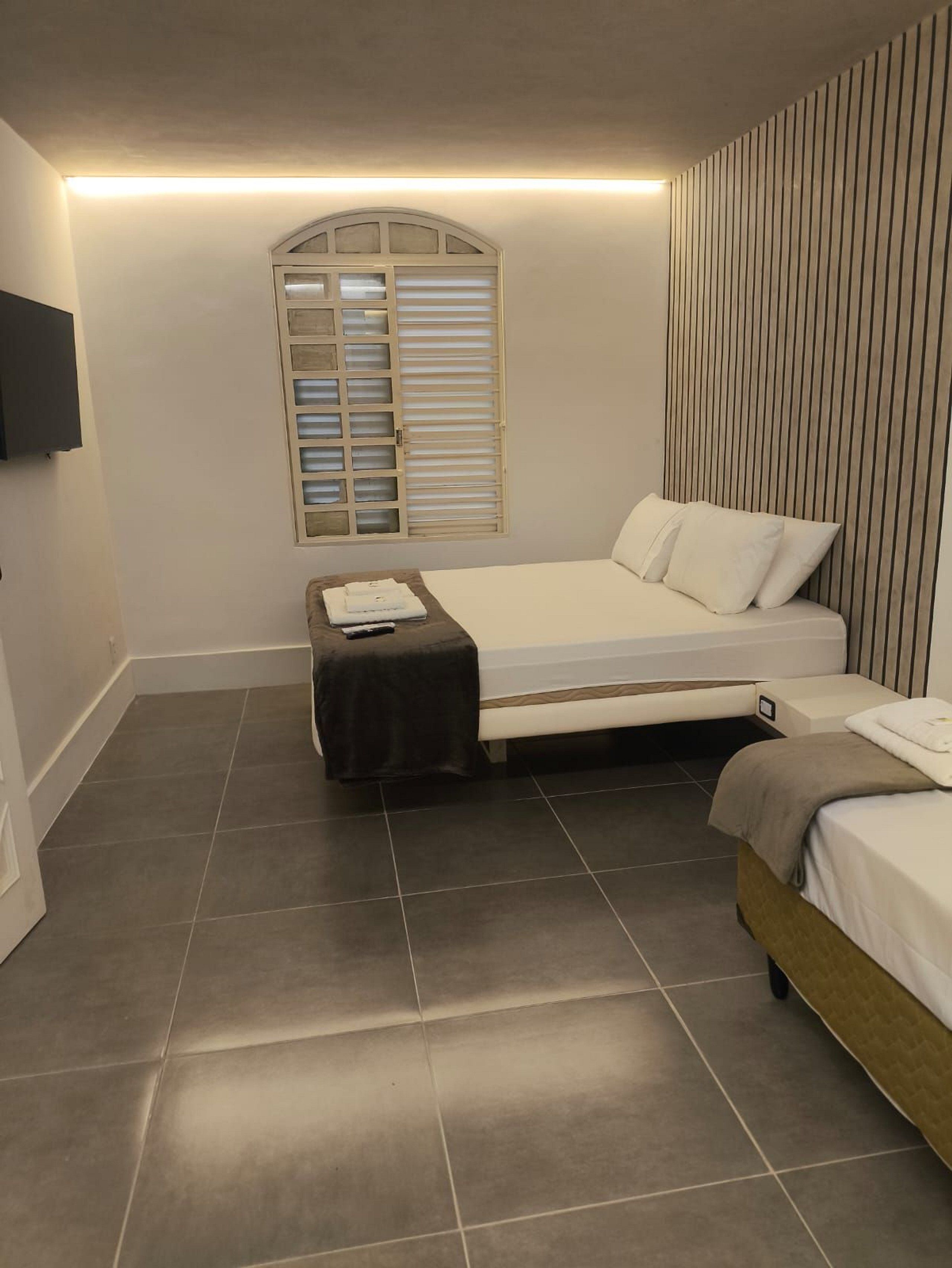 A bedroom with a bed, a bedside table, a window with blinds, and a wall-mounted TV.