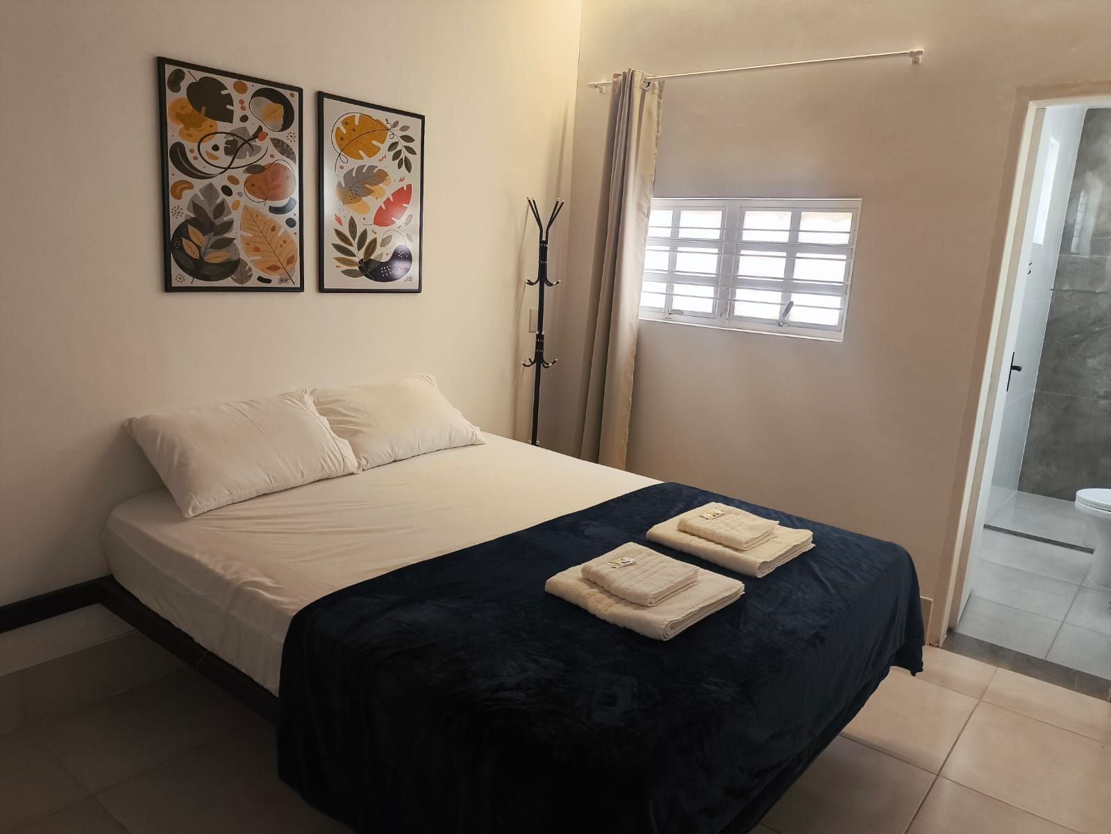 A bedroom with a bed, two framed artworks, a window with curtains, and a bathroom visible through an open door.