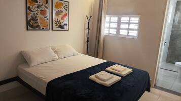 A bedroom with a bed, two framed artworks, a window with curtains, and a bathroom visible through an open door.