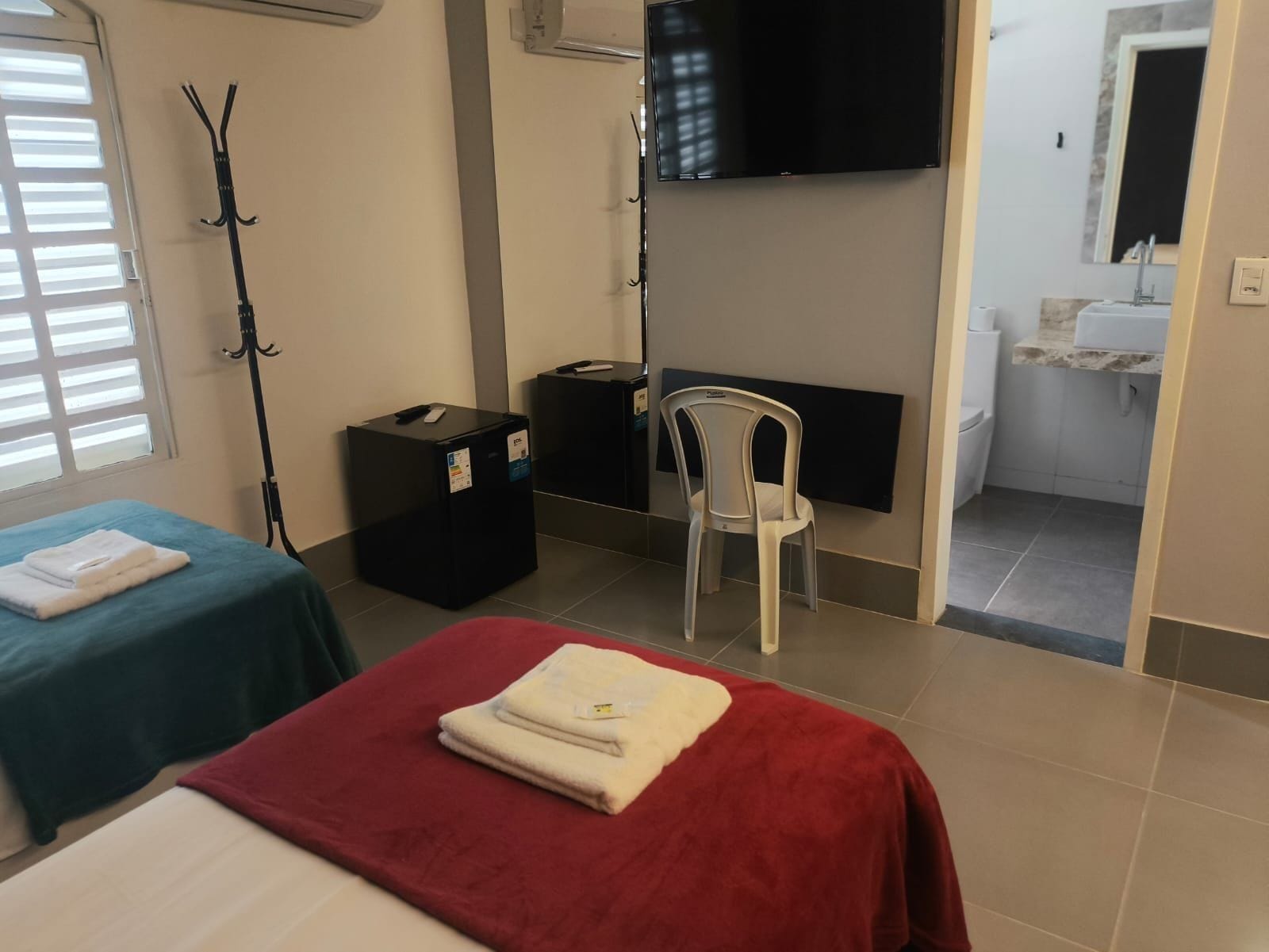 A hotel room with two beds, a TV, a chair, a small table, and a bathroom visible through an open door.