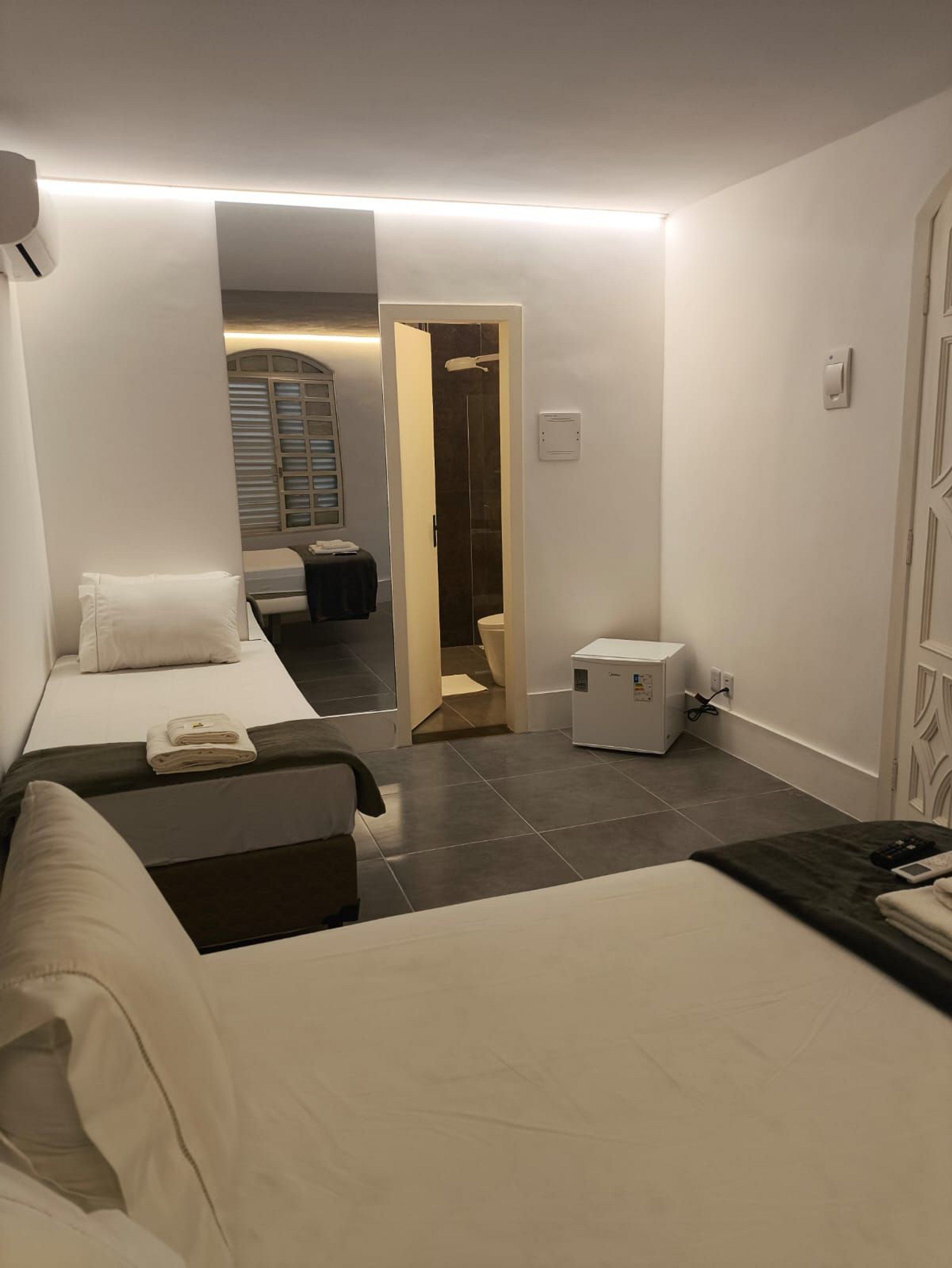 Quarto de hotel com cama, mesinha de cabeceira, escrivaninha e cadeira. Um banheiro é visível por uma porta aberta.