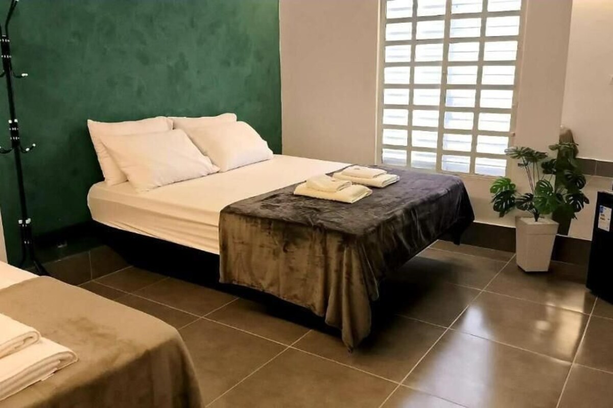 A hotel room with a bed, a small table with towels, a potted plant, and a desk with a computer.
