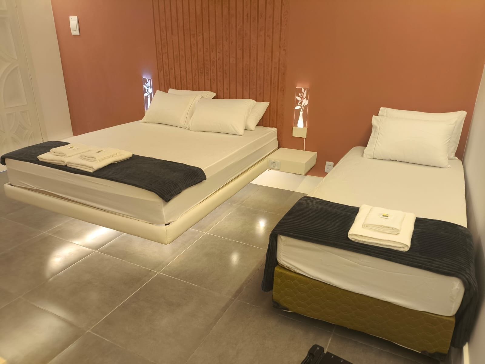 Uma cama de casal com cabeceira preta e uma cama simples com cabeceira de madeira em um quarto com piso de azulejos.