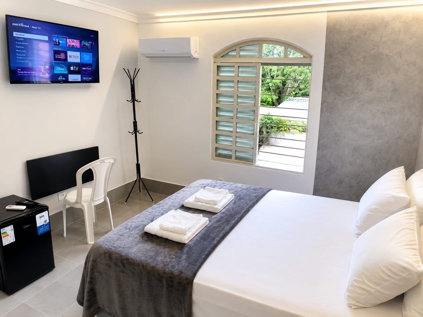 A hotel room with a bed, a desk, a chair, a TV displaying a channel list, an air conditioner, and a window with shutters.