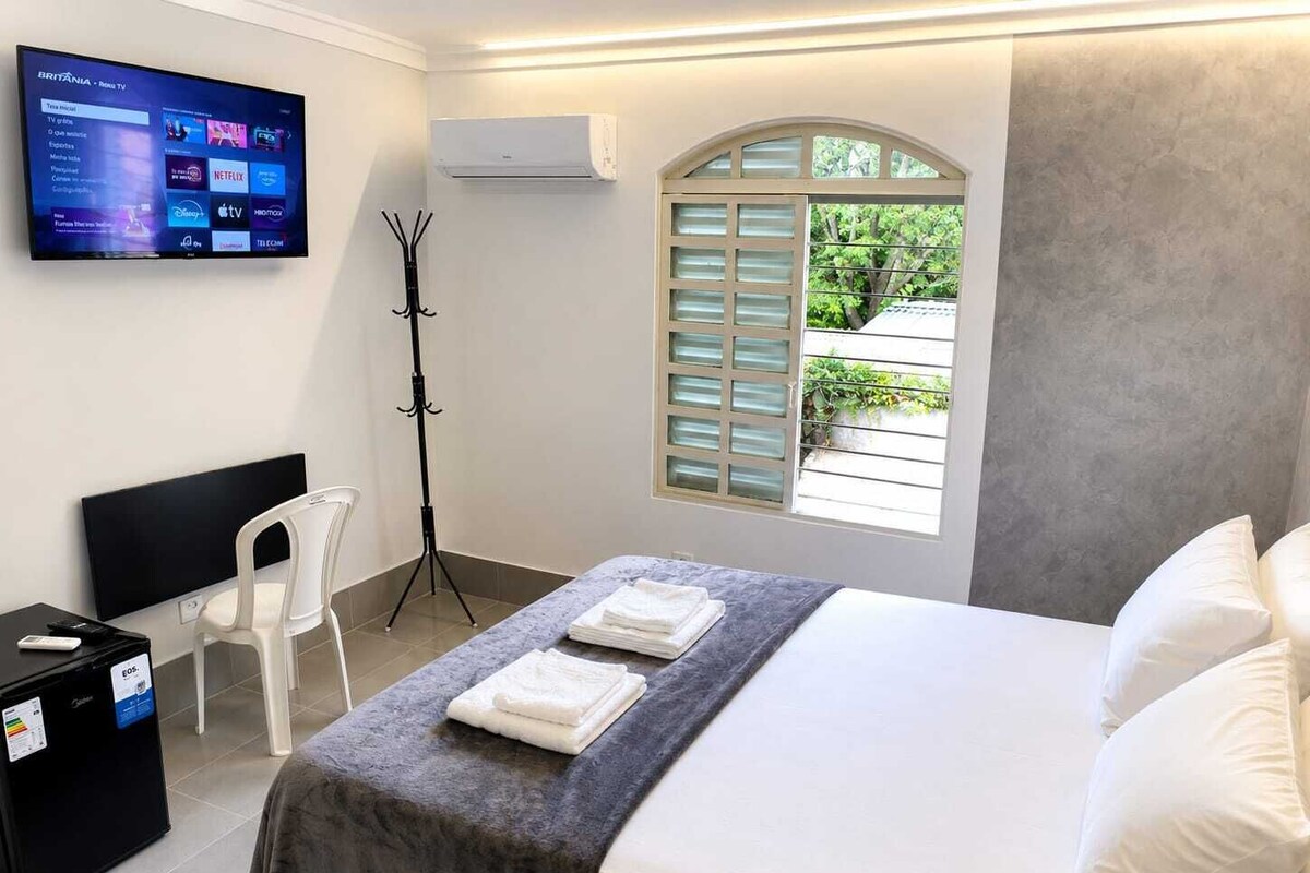 A hotel room with a bed, a desk, a chair, a TV displaying a channel list, an air conditioner, and a window with shutters.