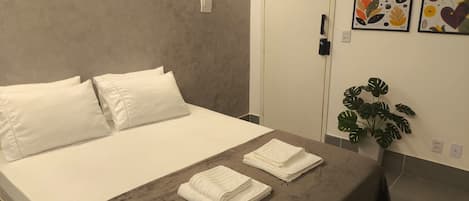 A hotel room with a bed, pillows, a bedside table with towels, a door, and a wall with artwork.