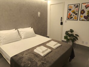 A hotel room with a bed, pillows, a bedside table with towels, a door, and a wall with artwork. - Gyn Suites Sul (Goiania)