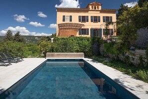 A swimming pool with a wooden deck, surrounded by greenery, and a large building with a tiled roof in the background. - Villa in Grasse with swimming pool (Grasse)
