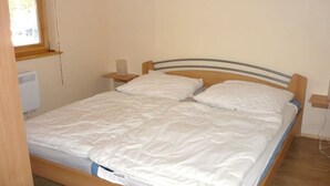 Cama de solteiro com cabeceira de madeira e roupa de cama branca em quarto com janela e aquecedor fixado na parede.