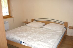 Un llit individual amb un panell de fusta al capçal i roba de llit blanca en una habitació amb finestra i un calefactor fixat a la paret.
