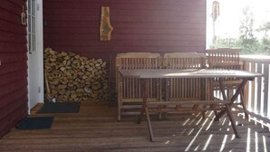 Um deck de madeira com mesa e cadeiras, uma pilha de lenha e uma porta para um edifício.
