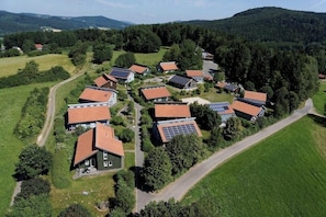 Eine Vogelperspektive auf eine Wohngegend mit Häusern, von denen einige mit Solaranlagen ausgestattet sind, umgeben von viel Grün und Hügeln.