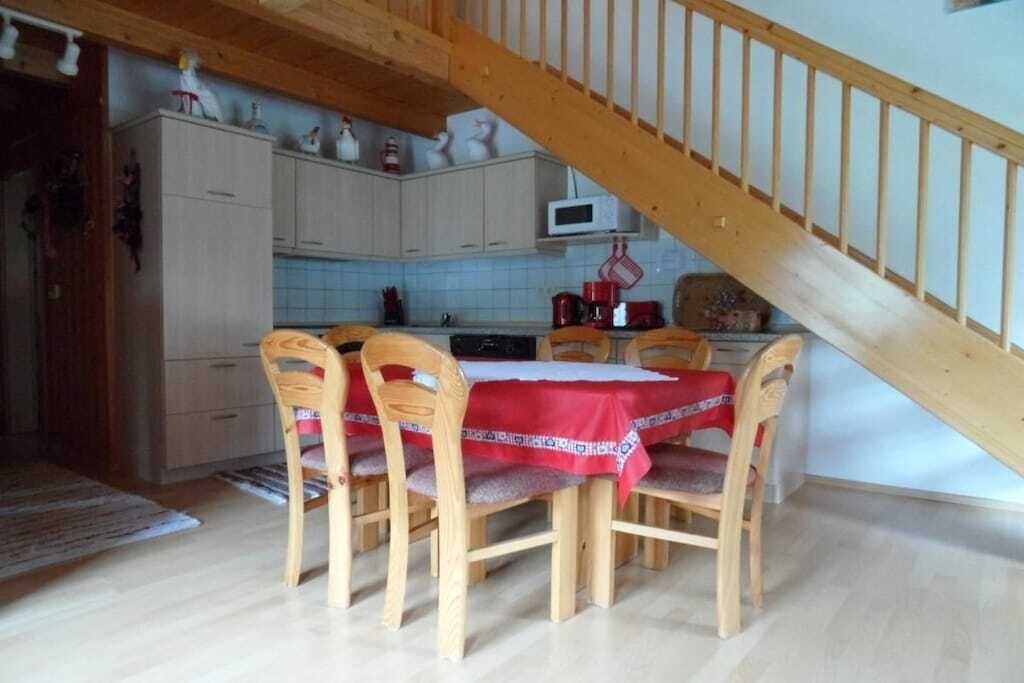 Un coin repas avec une table et des chaises en bois, une cuisine à l’arrière-plan et un escalier en bois.