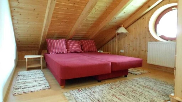 Une petite pièce chaleureuse avec un plafond en bois, un lit avec une literie rouge, une petite table et une fenêtre ronde.