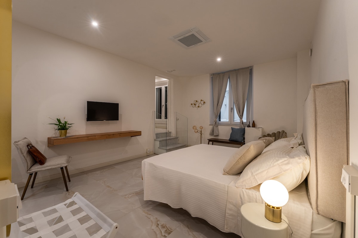 A bedroom with a bed, a bedside table with a lamp, a TV mounted on the wall, and a window with curtains.