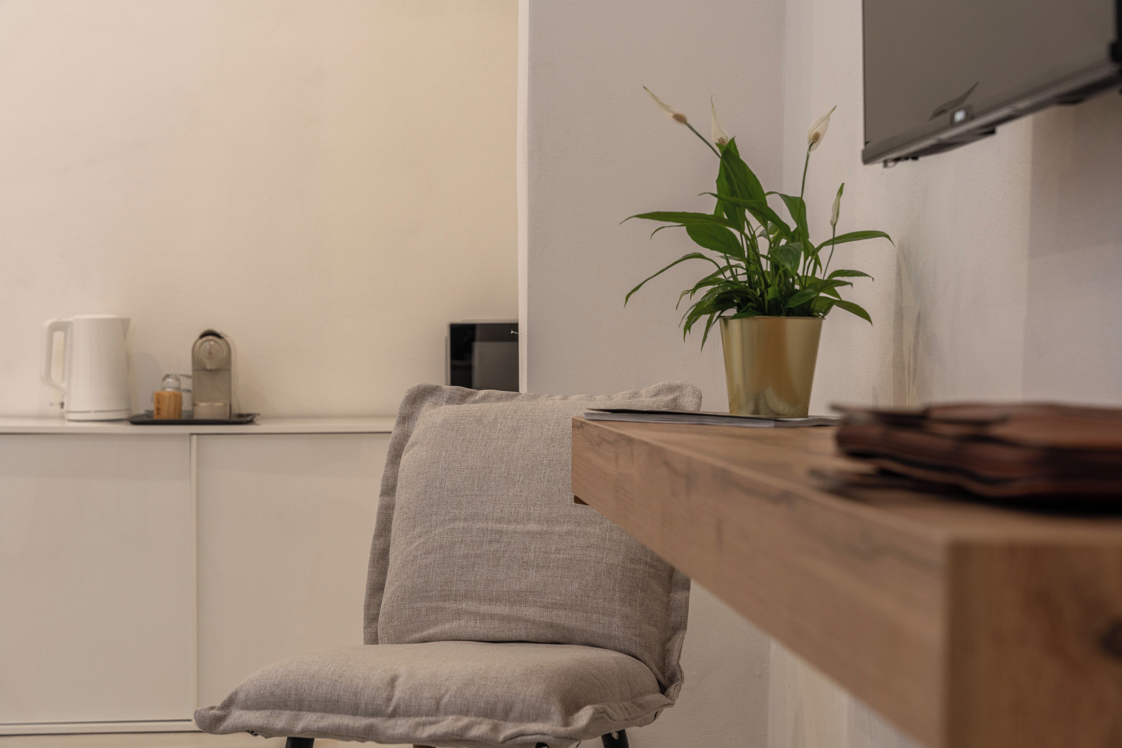 Une pièce au décor minimaliste, avec un bureau en bois, une chaise grise, une plante en pot, un téléviseur à écran plat fixé au mur et une machine à café posée sur le comptoir.