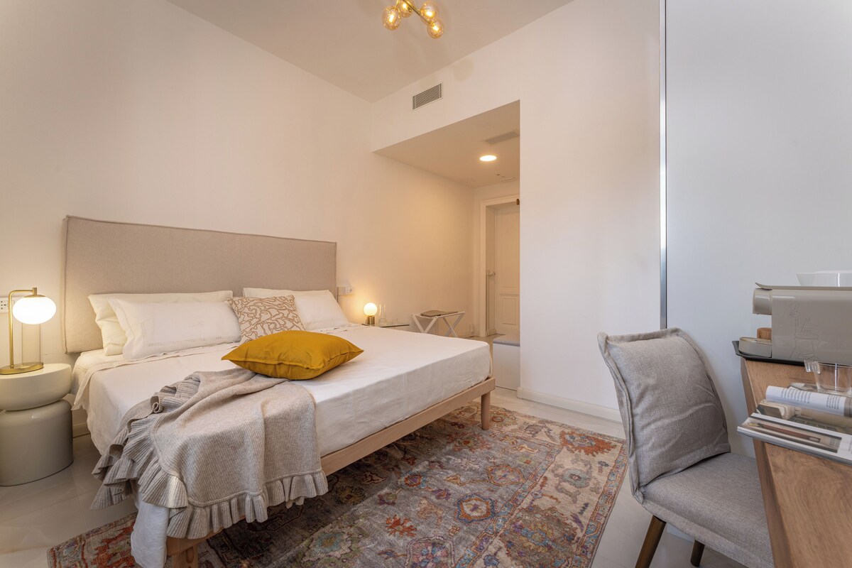 A modern bedroom with a bed, bedside lamps, a chair, and a desk with a coffee machine.