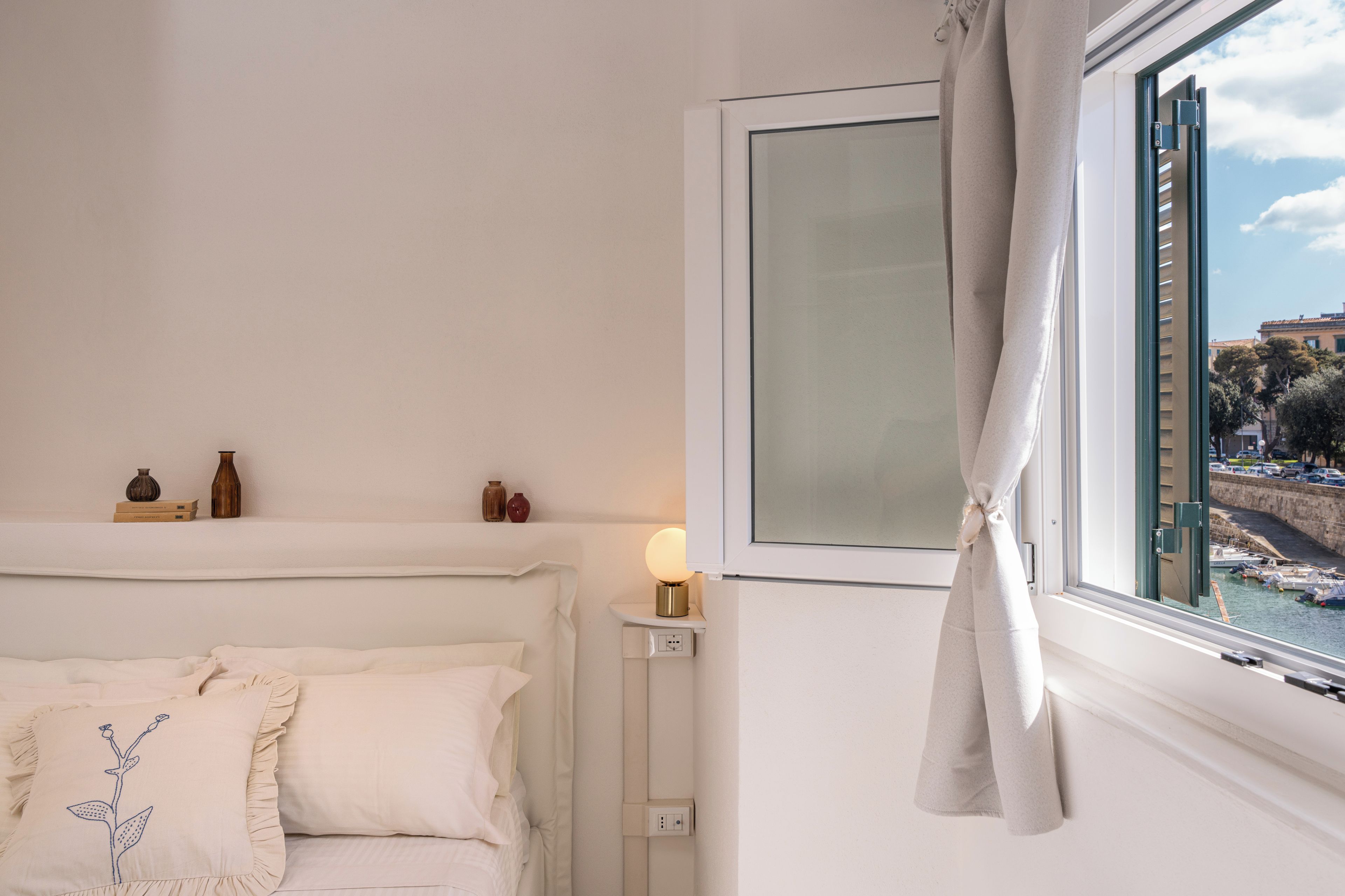 Quarto com cama, mesa de cabeceira, abajur e janela com vista para um porto.