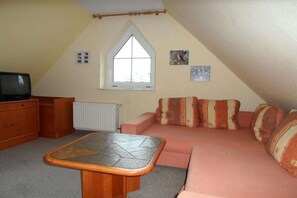 Ein gemütliches Schlafzimmer im Dachgeschoss mit schrägen Wänden, einem Fenster, einer Couch mit gemusterten Kissen, einem kleinen Tisch und einem Fernseher.