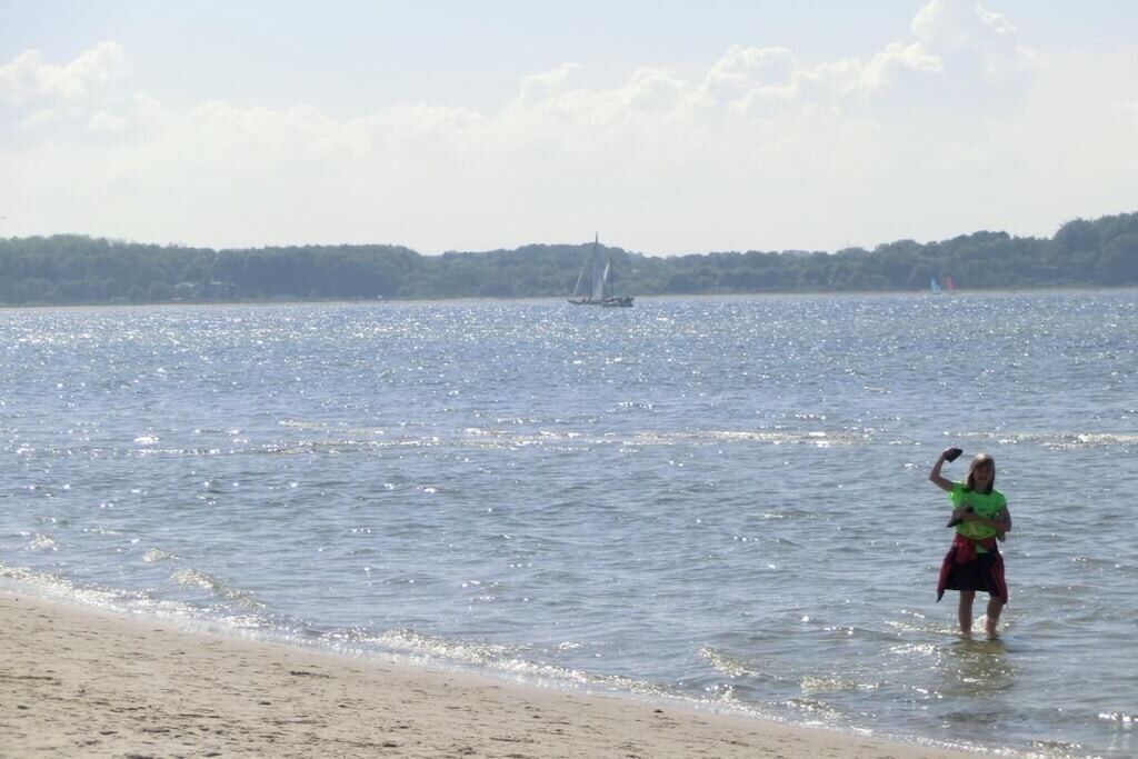 A beach with a person standing in shallow water, a sailboat in the distance, and a forested shoreline.