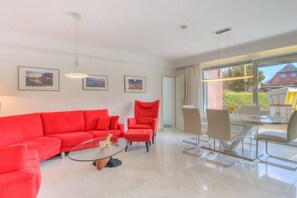 Een moderne woonkamer met een rode bank, een glazen salontafel en een eethoek met witte stoelen.