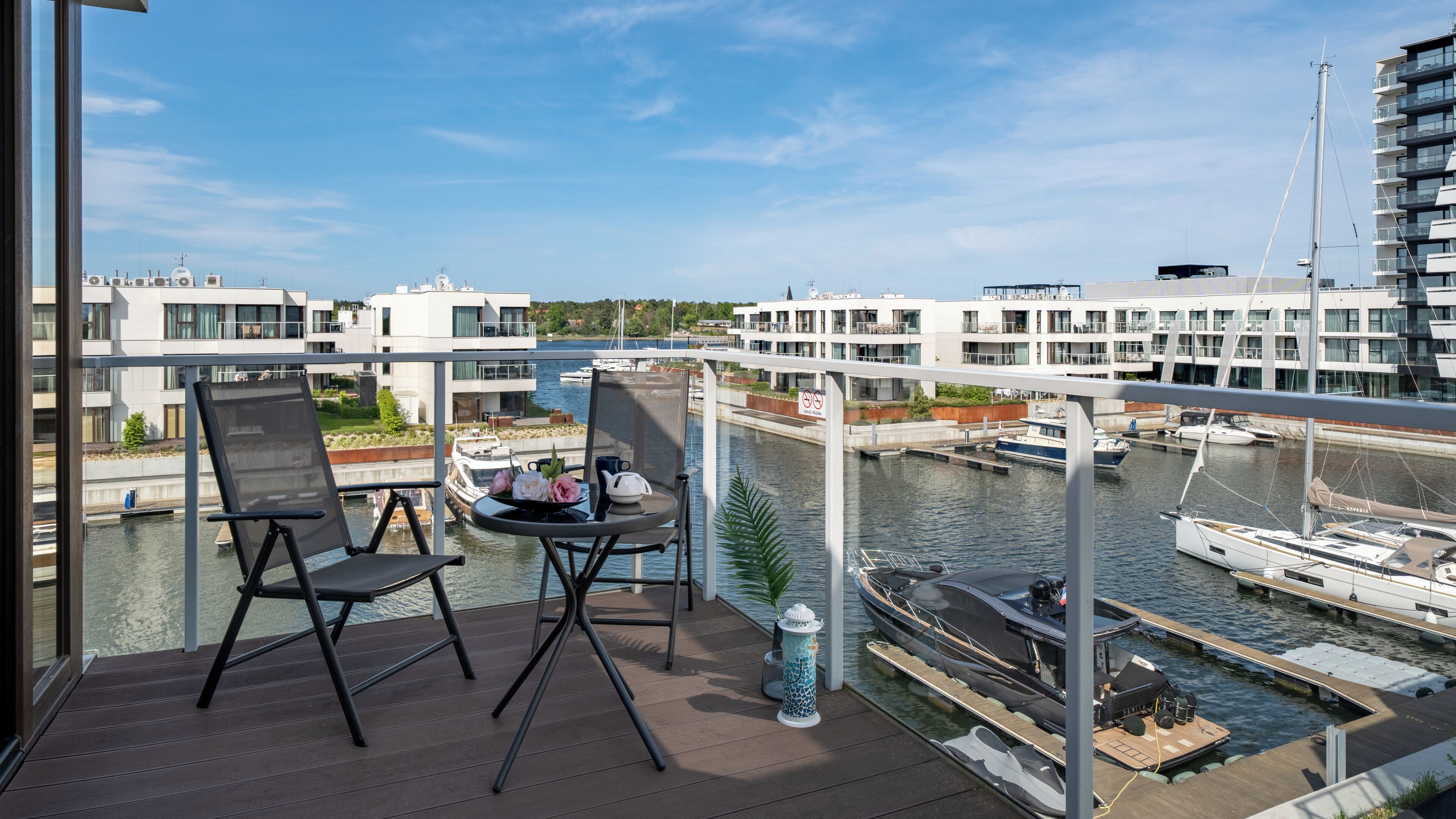 A balcony with two chairs and a table, overlooking a marina with boats and modern buildings.