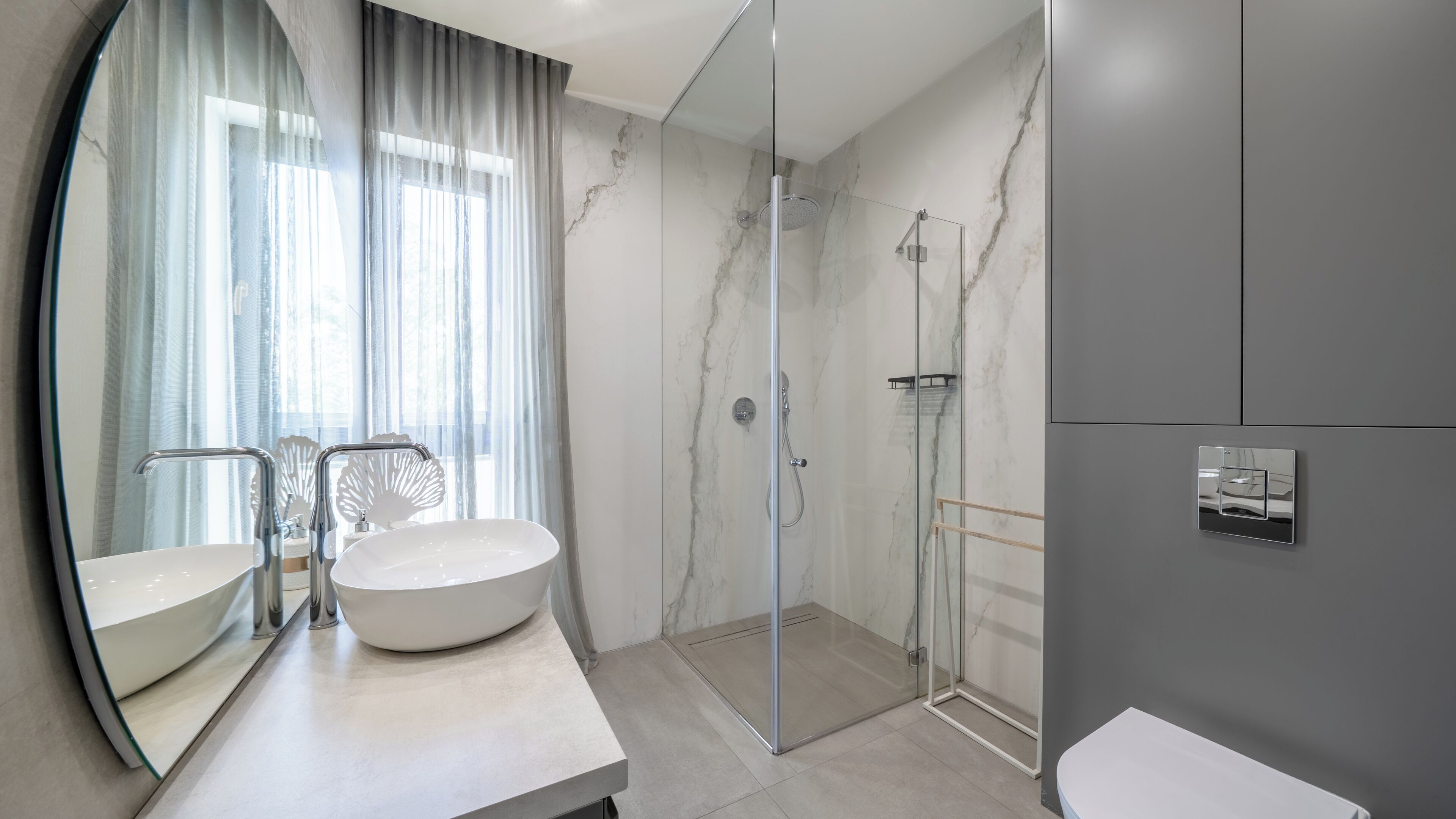 Un baño moderno con un espejo grande, un lavabo redondo, ducha con mampara de vidrio y pared de mármol.