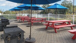 Outdoor picnic tables with blue umbrellas, a grill, and a white fence. - Myrtle Beach | Beach Escape | Pool & Boardwalk | Hotel (Myrtle Beach)