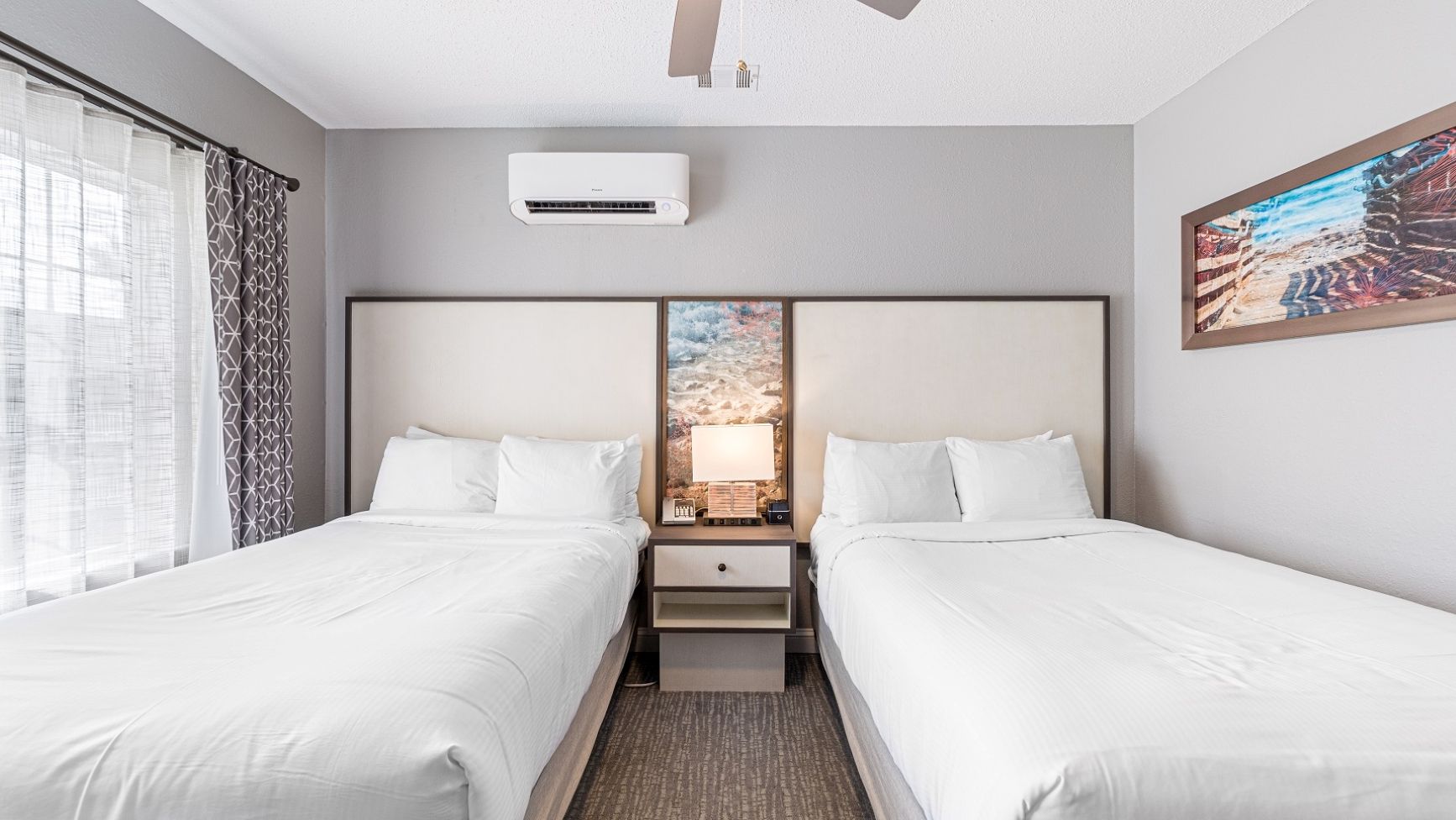 Quarto de hotel com duas camas, uma mesa de cabeceira, uma luminária, um aparelho de ar condicionado e um quadro na parede.