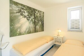 A single bed with a white mattress and a yellow blanket, a bedside table with a lamp, and a large forest-themed painting on the wall. - Holiday apartment in Sylt with 1 bedroom (Wenningstedt)